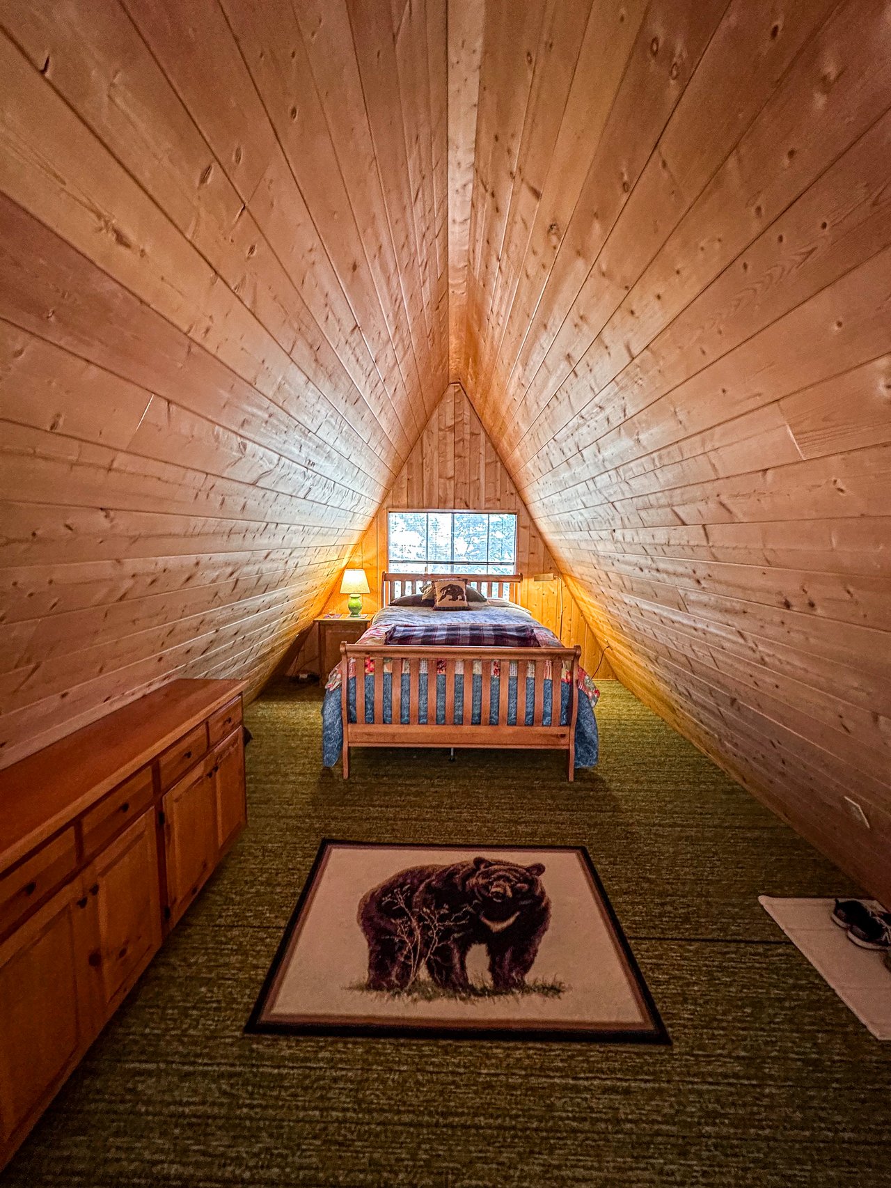 Furnished A-frame bedroom with a wooden bed, plank walls, and a bear-patterned rug.