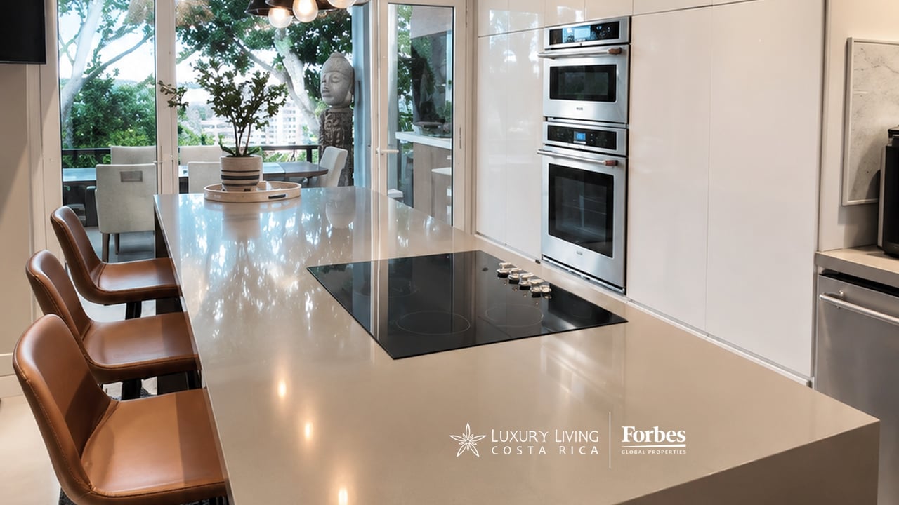 Modern kitchen with a long stone island, black cooktop, and built-in stainless-steel ovens beside glass doors.