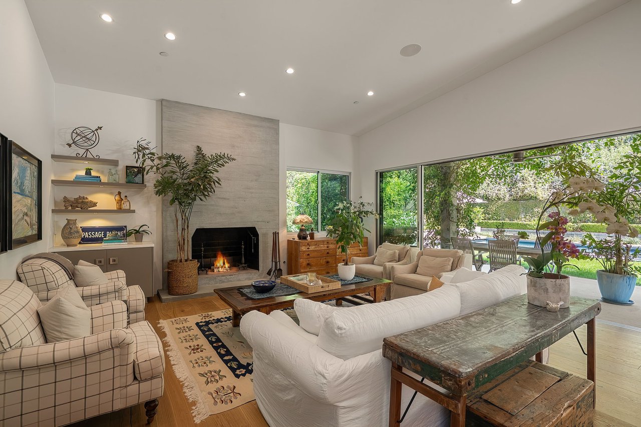 Living room with a stone fireplace, white sofas, and glass doors overlooking the garden and pool.
