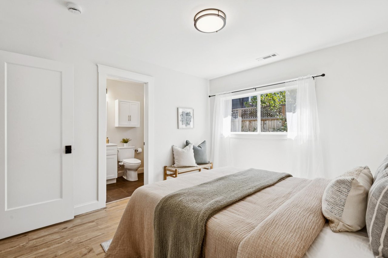 Bright bedroom with neutral bedding, a curtained window, and an open doorway to the ensuite bathroom.