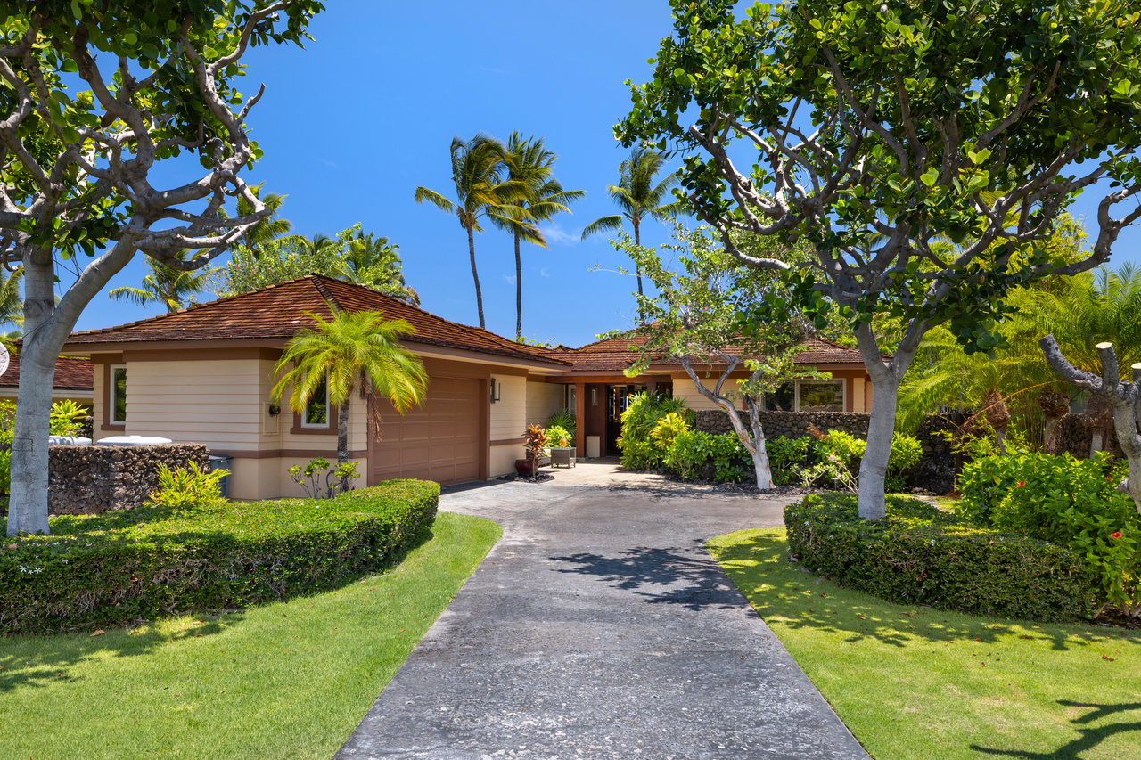 Single-story beige home with a tiled roof, curved driveway, palm trees, and tropical landscaping.
