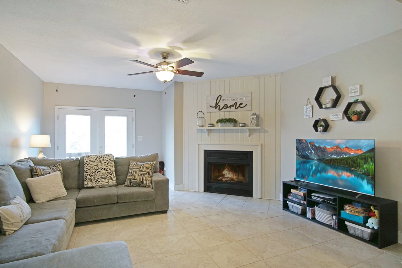 A cozy living room with a gray sectional sofa, patterned cushions, a lit fireplace under a "Home" sign, and a large TV. Soft lighting creates a warm ambiance.