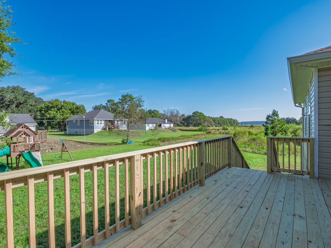 Wooden backyard deck overlooking a lawn, children's playset, neighboring homes, and distant water.