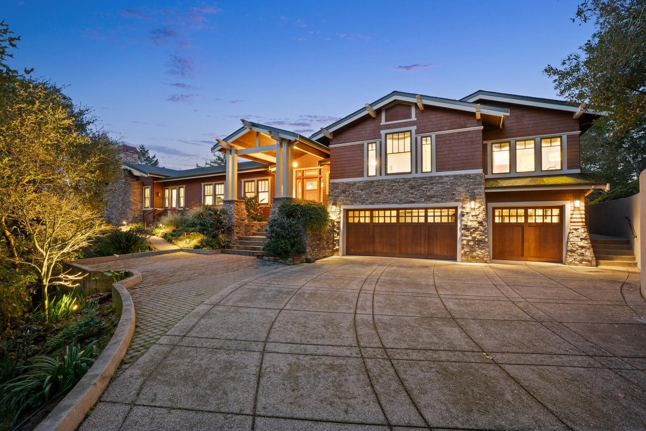 Two-story stone-and-shingle home with illuminated windows, two garages, and a landscaped driveway at dusk.