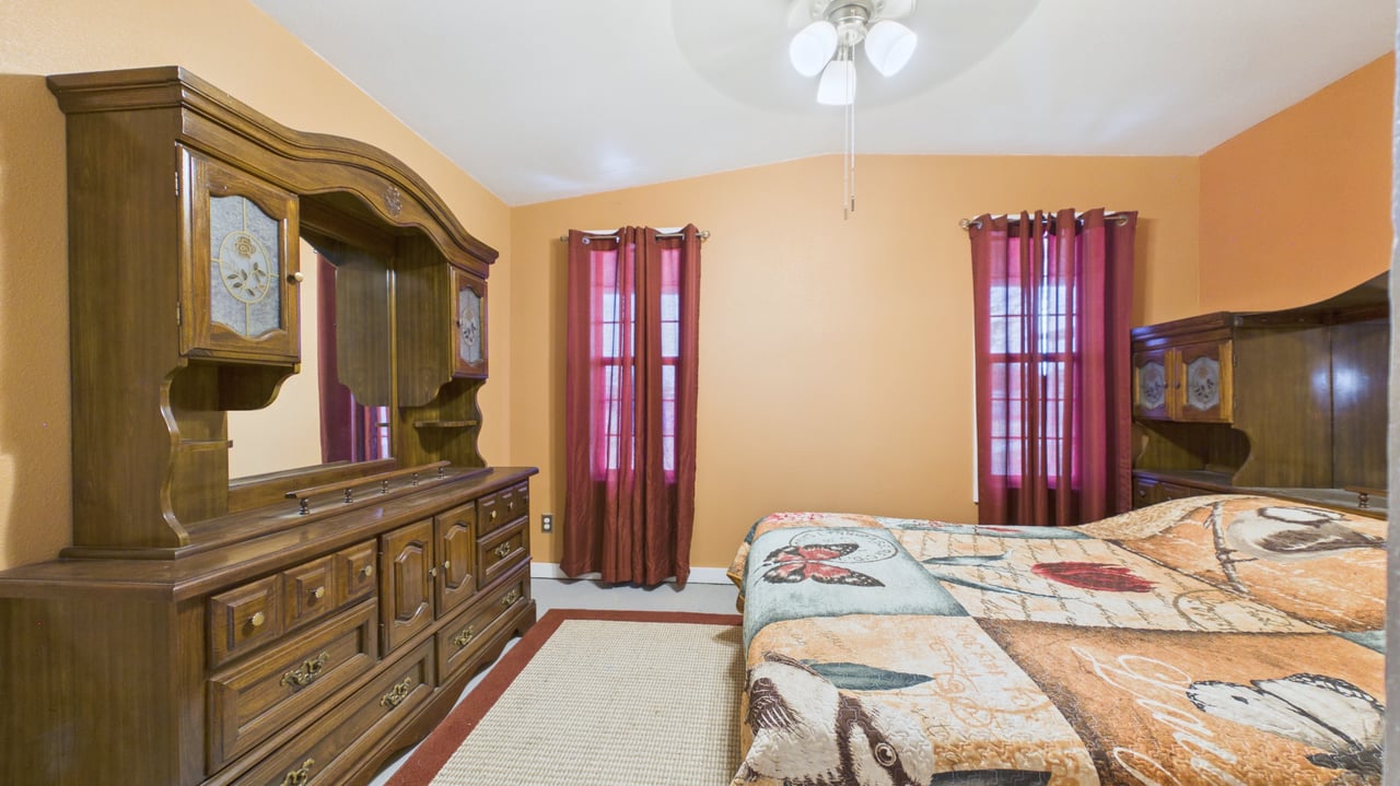 Bedroom with a patterned bedspread, large dark wood dresser and mirror, and burgundy curtains.