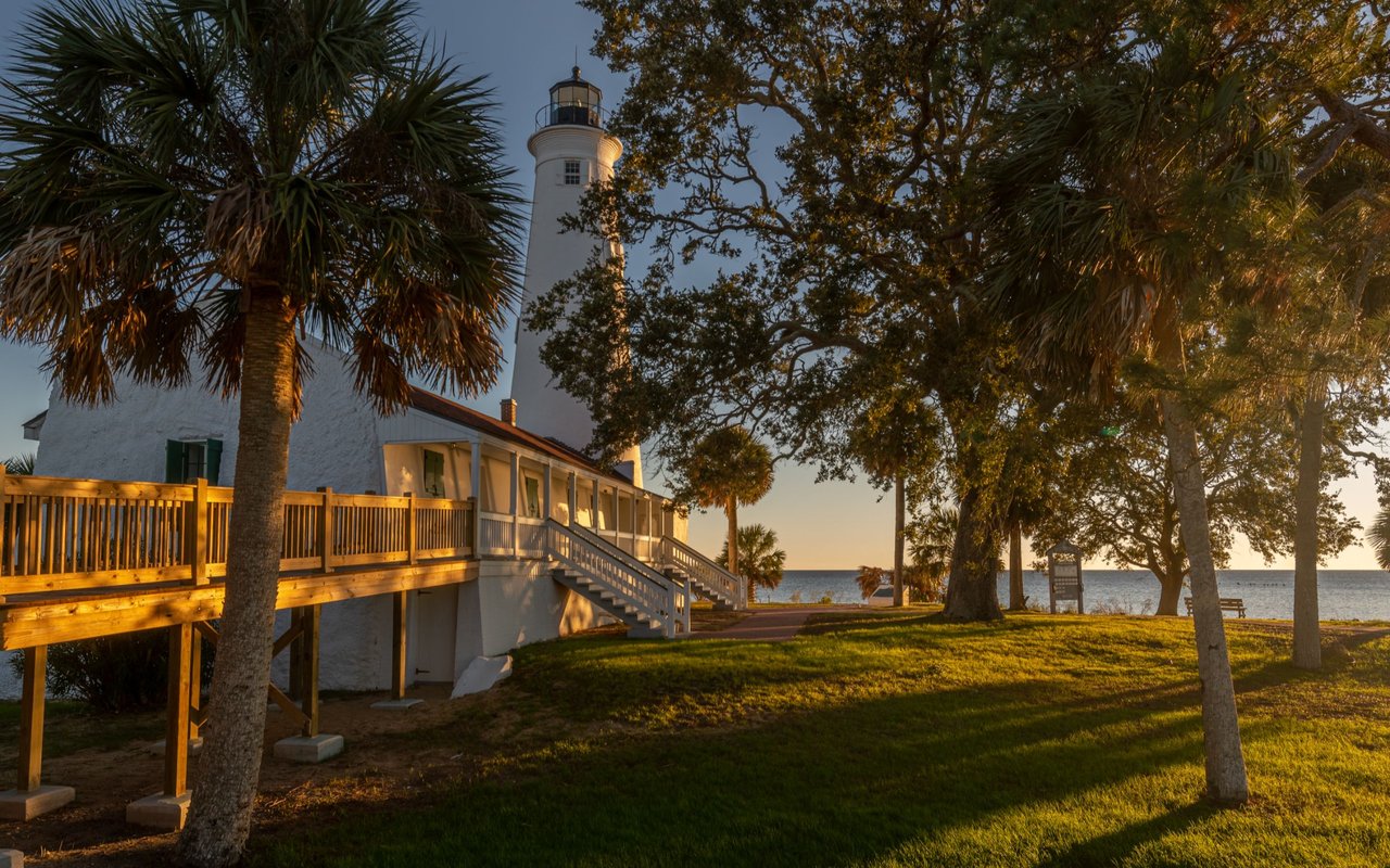 White lighthouse and waterfront building framed by trees beside the water at golden hour.