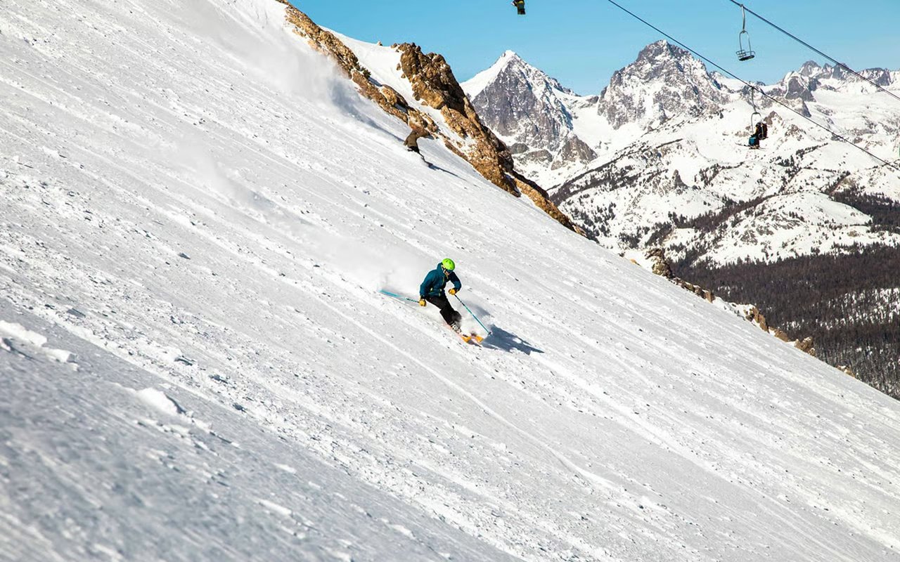 A skier descends a snowy mountain slope beneath rugged peaks and chairlifts.