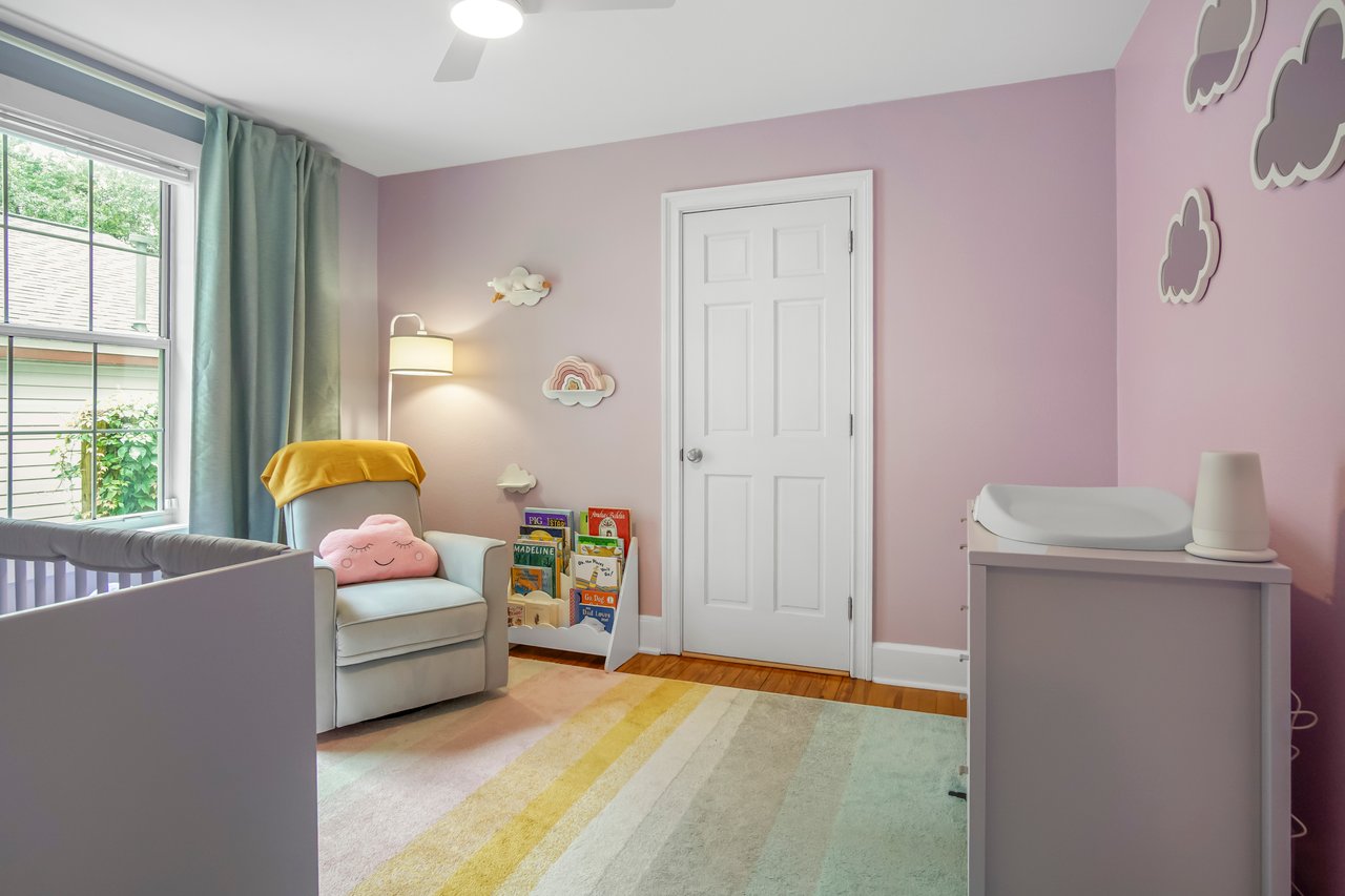 A pastel nursery with pink walls, gray crib, and changing table. An armchair with a yellow blanket sits by a window with green curtains. Soft, cozy ambiance.