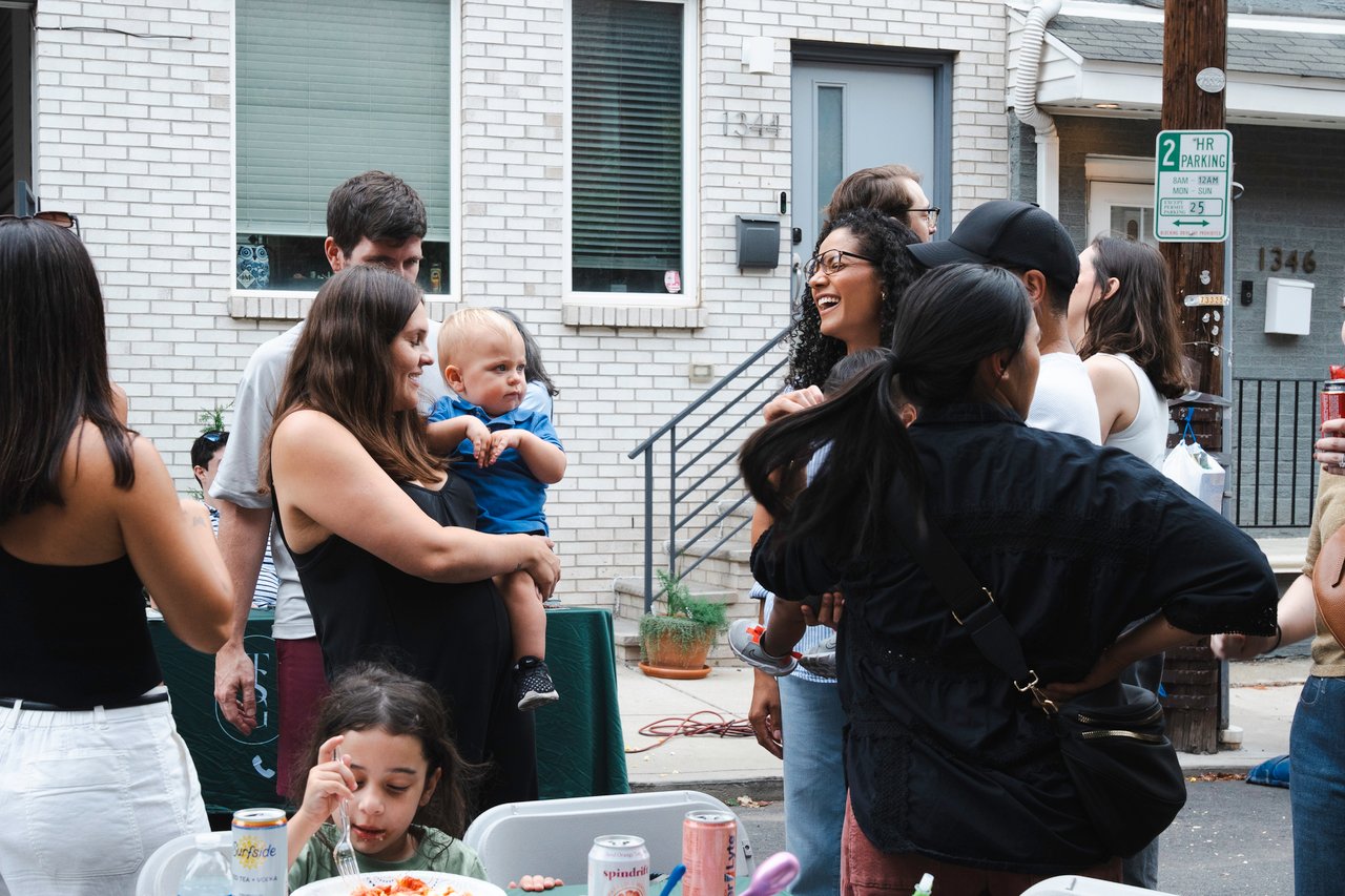 Neighbors mingle at an outdoor block party as a woman holds a baby and a child eats at a table.