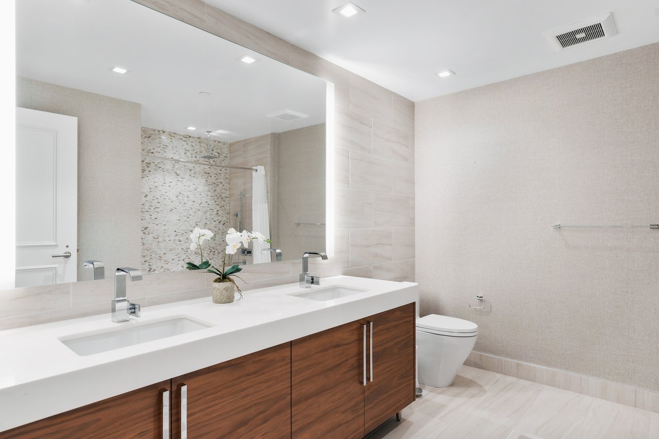 Modern bathroom with a double-sink dark wood vanity, illuminated mirror, and mosaic shower wall.