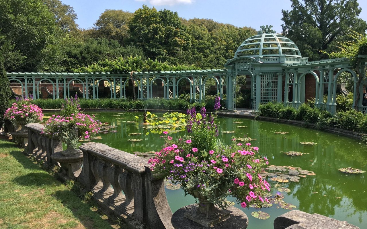 Formal garden pond with lily pads, flower-filled urns, and a pale green domed pergola.
