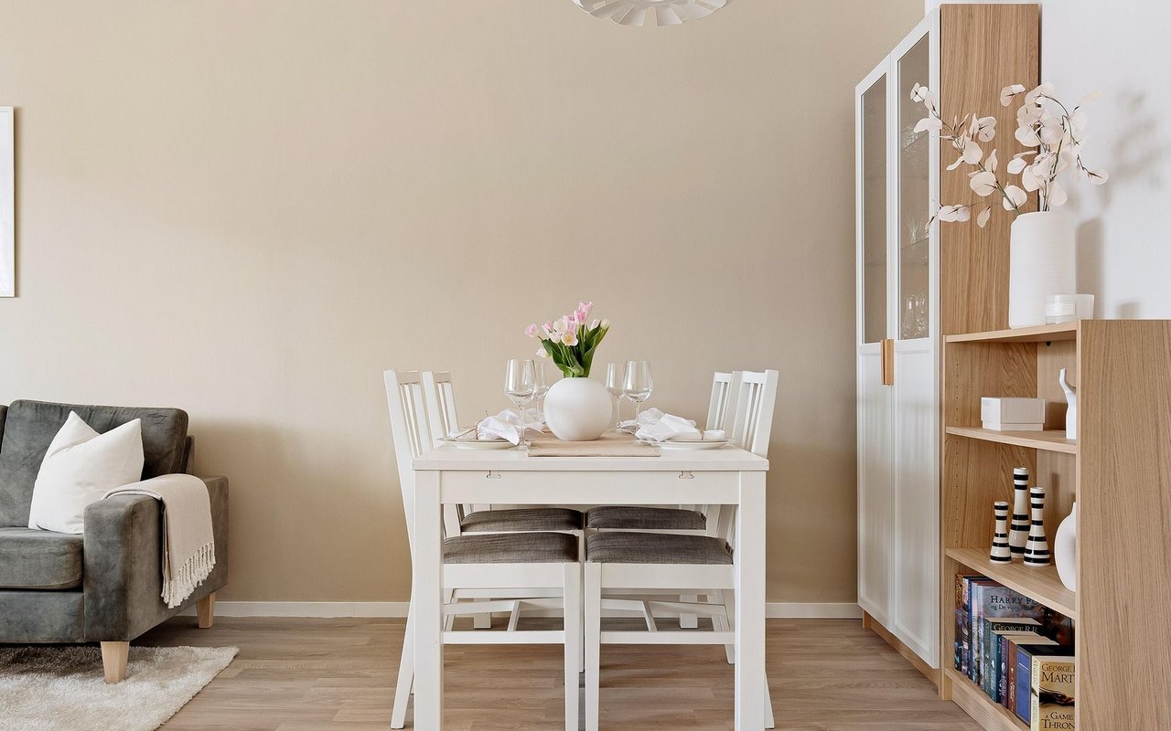 Dining area with a white table set for four, pink flowers, and light wood shelving beside a gray sofa.