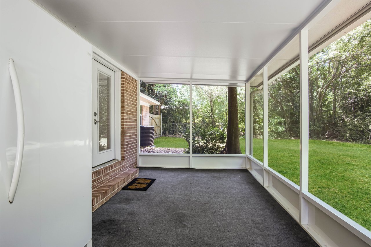 Enclosed porch with a glass door and windows, overlooking a lush green yard. A serene, bright space with a brick wall and dark carpeted floor.