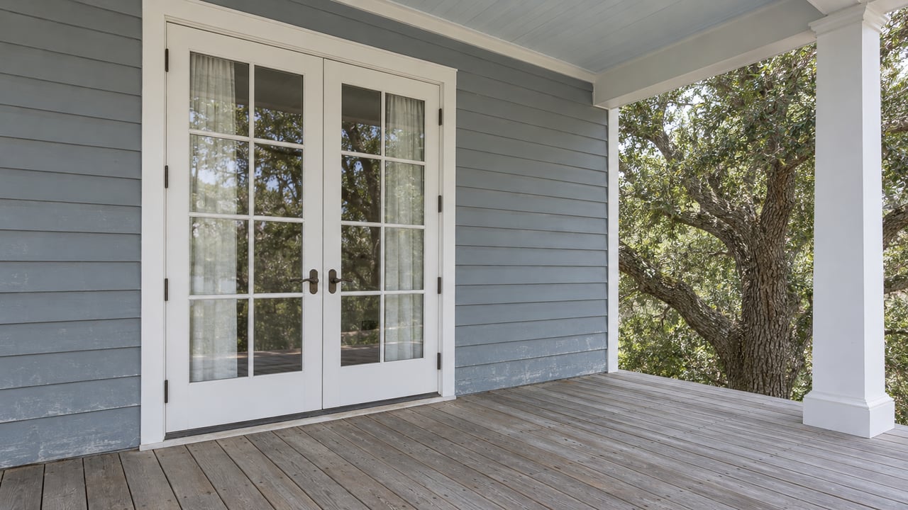 Gray-blue clapboard and closed white French doors on a weathered covered porch, with a mature live oak beyond a white column.
