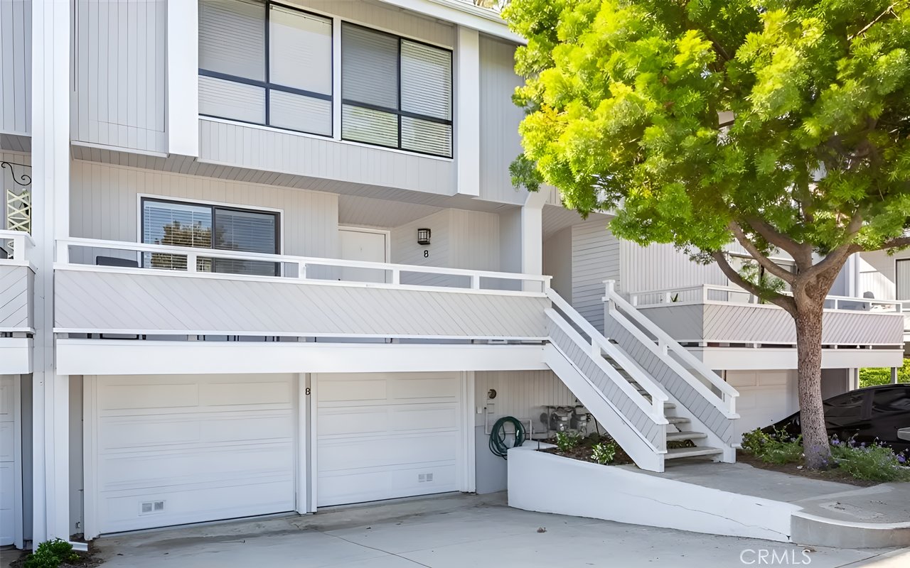 A modern gray two-story condominium with two white garage doors, a white staircase, and a large tree in front.