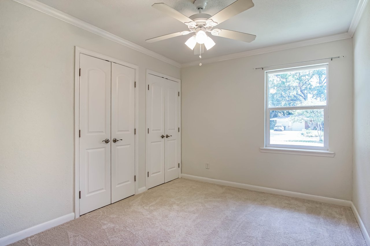 Empty room with beige carpet, white walls, and a ceiling fan with light. Two white closet doors are on the left, and a window on the right shows trees outside.