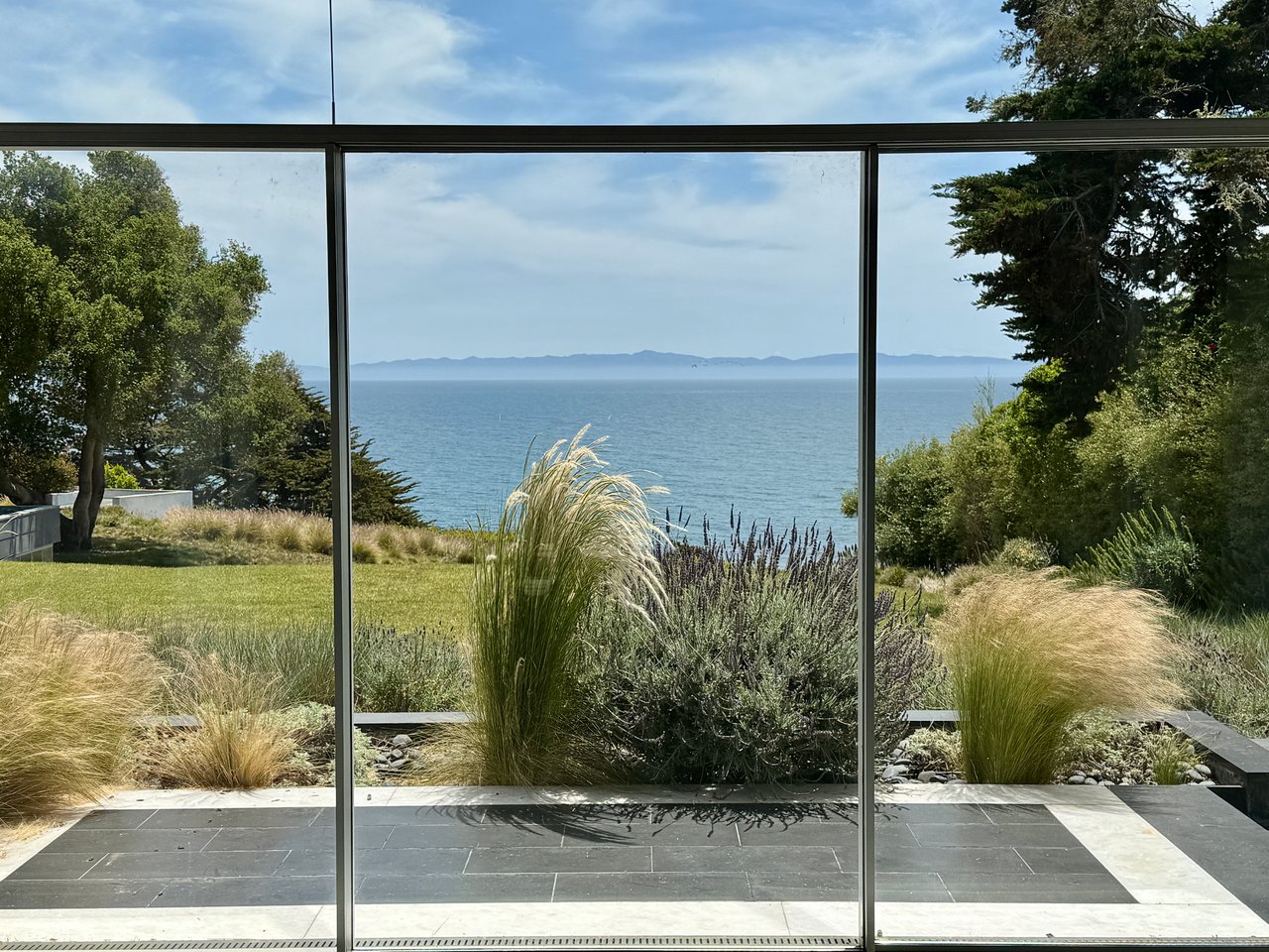 Ocean view through glass doors framed by drought-tolerant grasses and lavender landscaping, with the Channel Islands visible on the horizon.