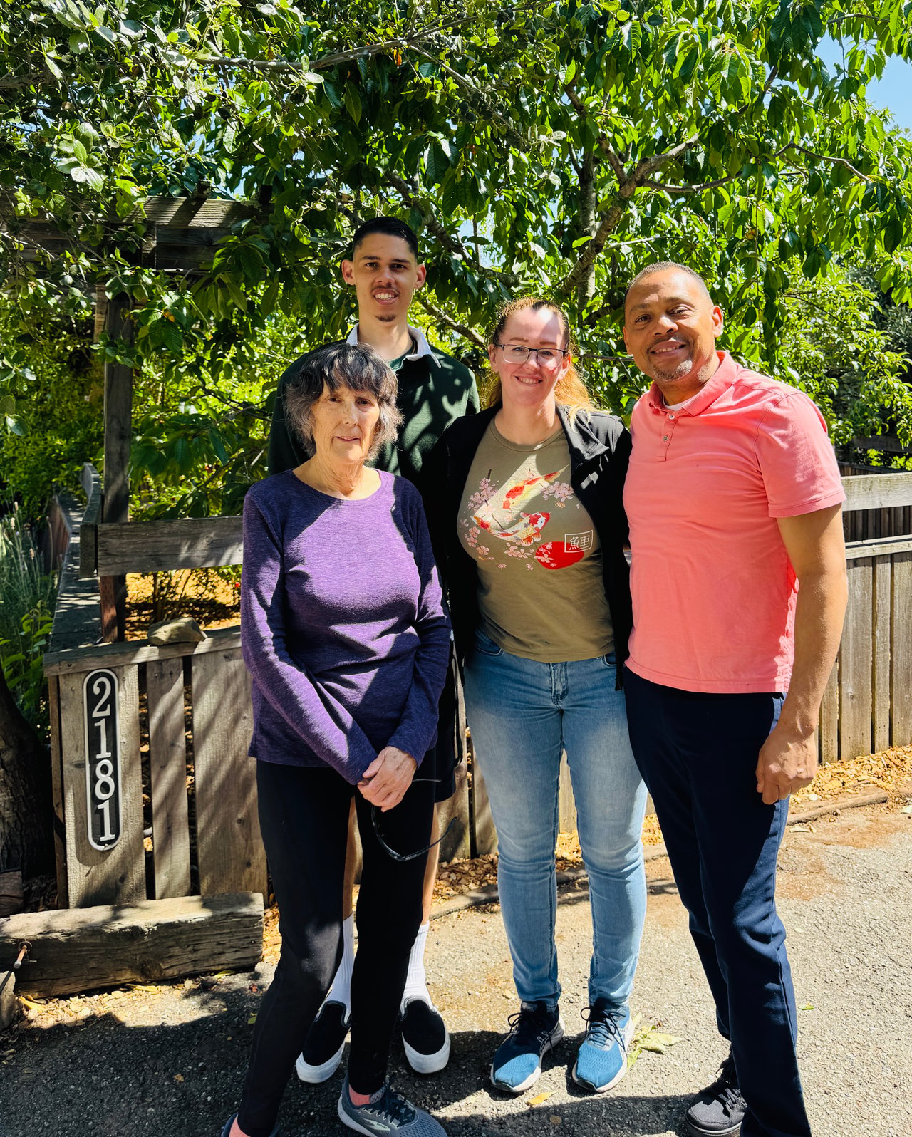 Four people pose together outdoors beside a wooden fence marked 2181.