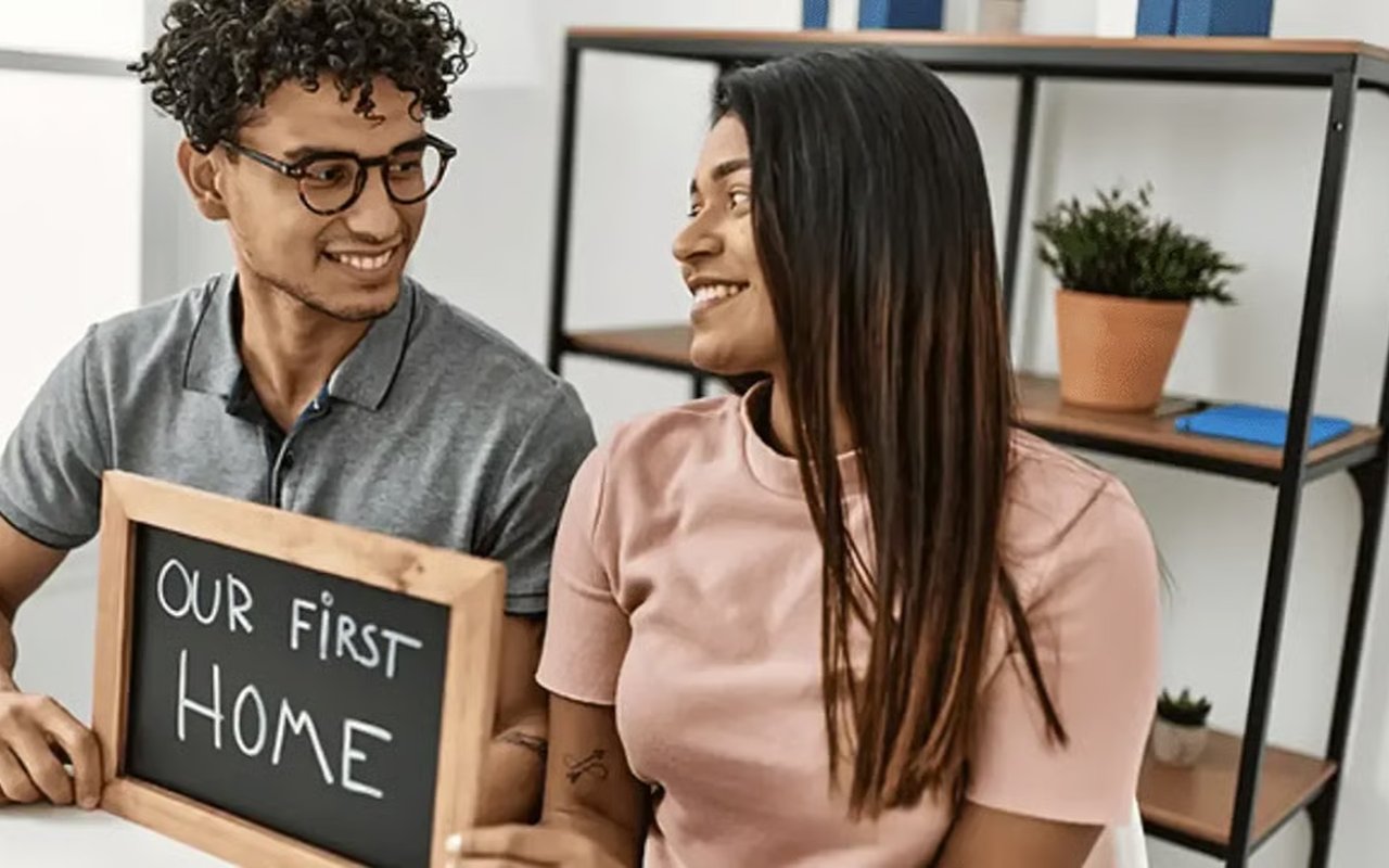Smiling couple holding a chalkboard that reads “OUR FIRST HOME.”