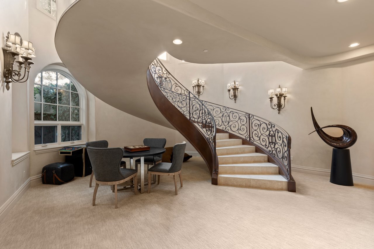 Foyer with a sweeping curved staircase, ornate iron railing, arched window, and small seating area.