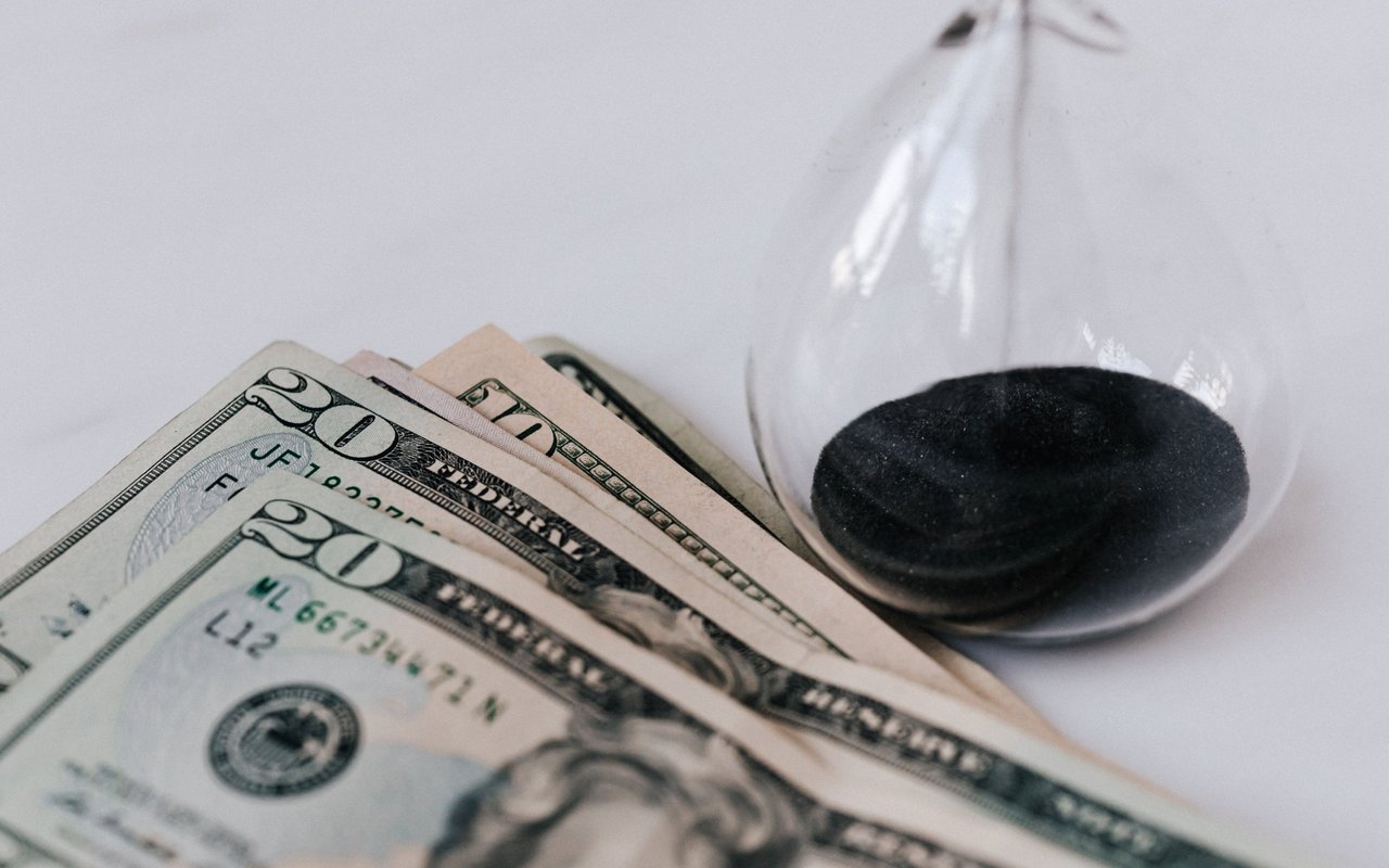 U.S. dollar bills beside a glass hourglass filled with dark sand.