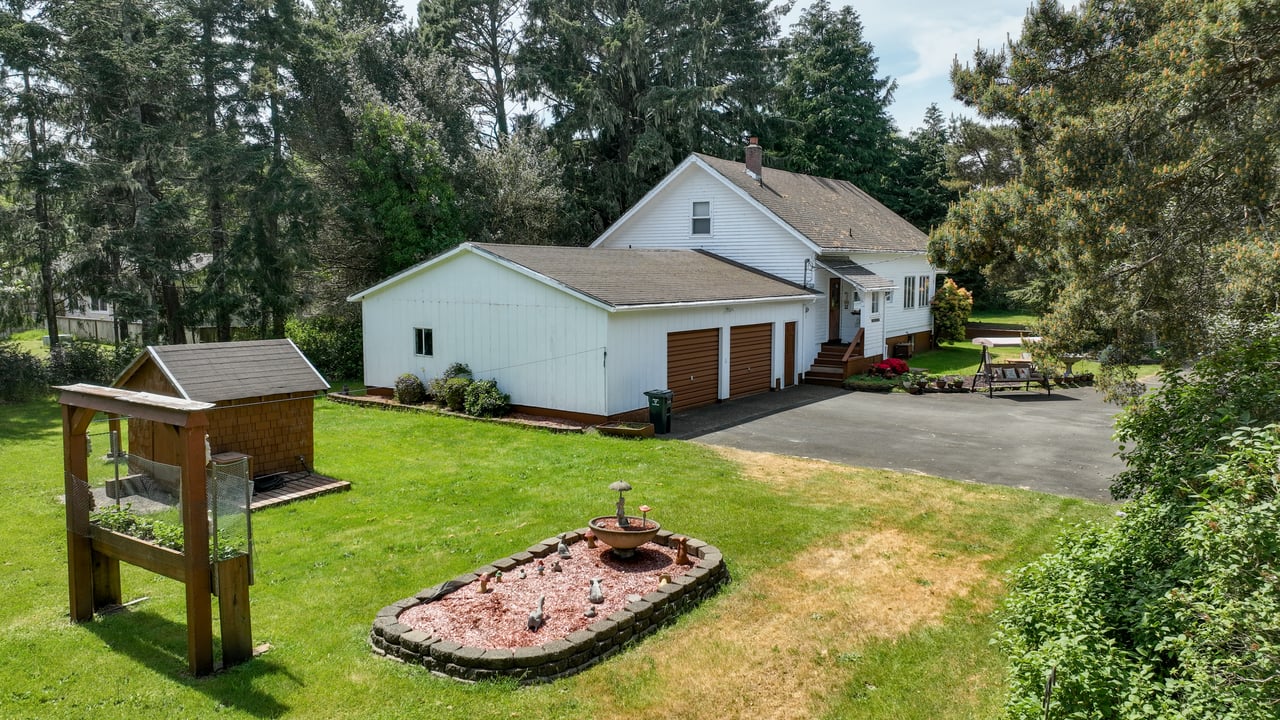 Spacious white home with a detached garage, large green backyard, mature evergreen trees, and a circular fire pit area with a birdbath.