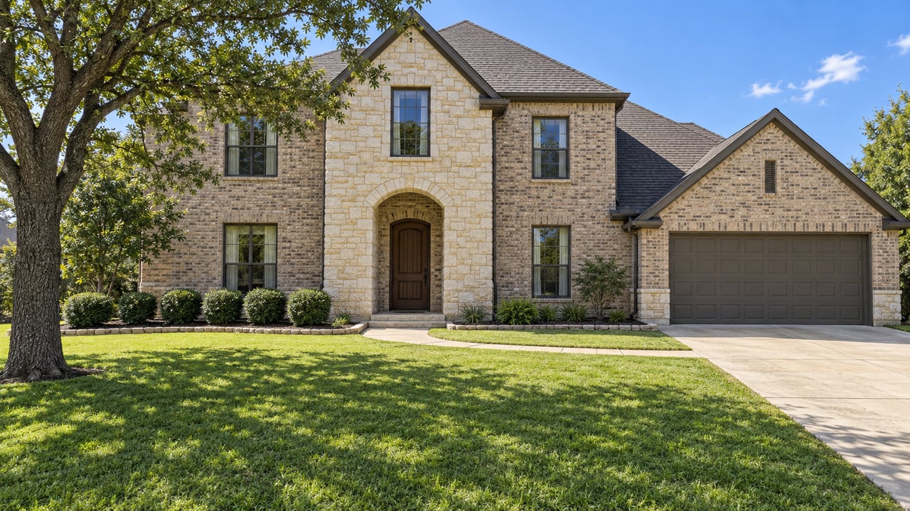 Two-story limestone and buff-brick home with dark-framed windows, a recessed arched entrance, broad lawn and a mature oak.