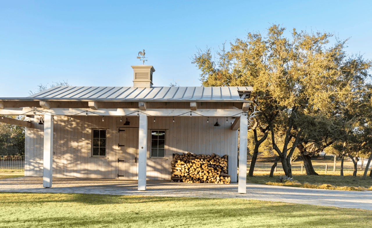 Ranch outbuilding with a metal roof, stacked firewood, and oak trees in golden light.