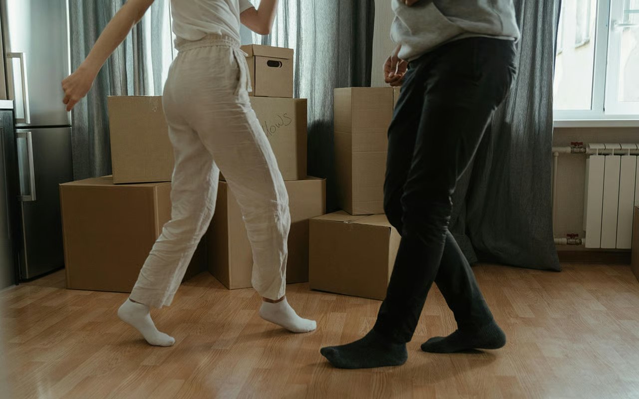 A couple walks among cardboard moving boxes in a bright, curtained living room.