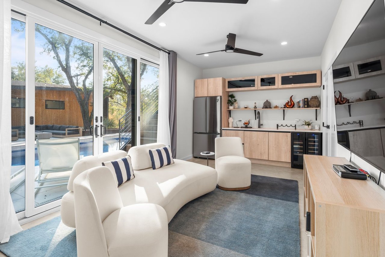 Living room with curved white seating and a built-in kitchenette beside sliding doors overlooking a pool.