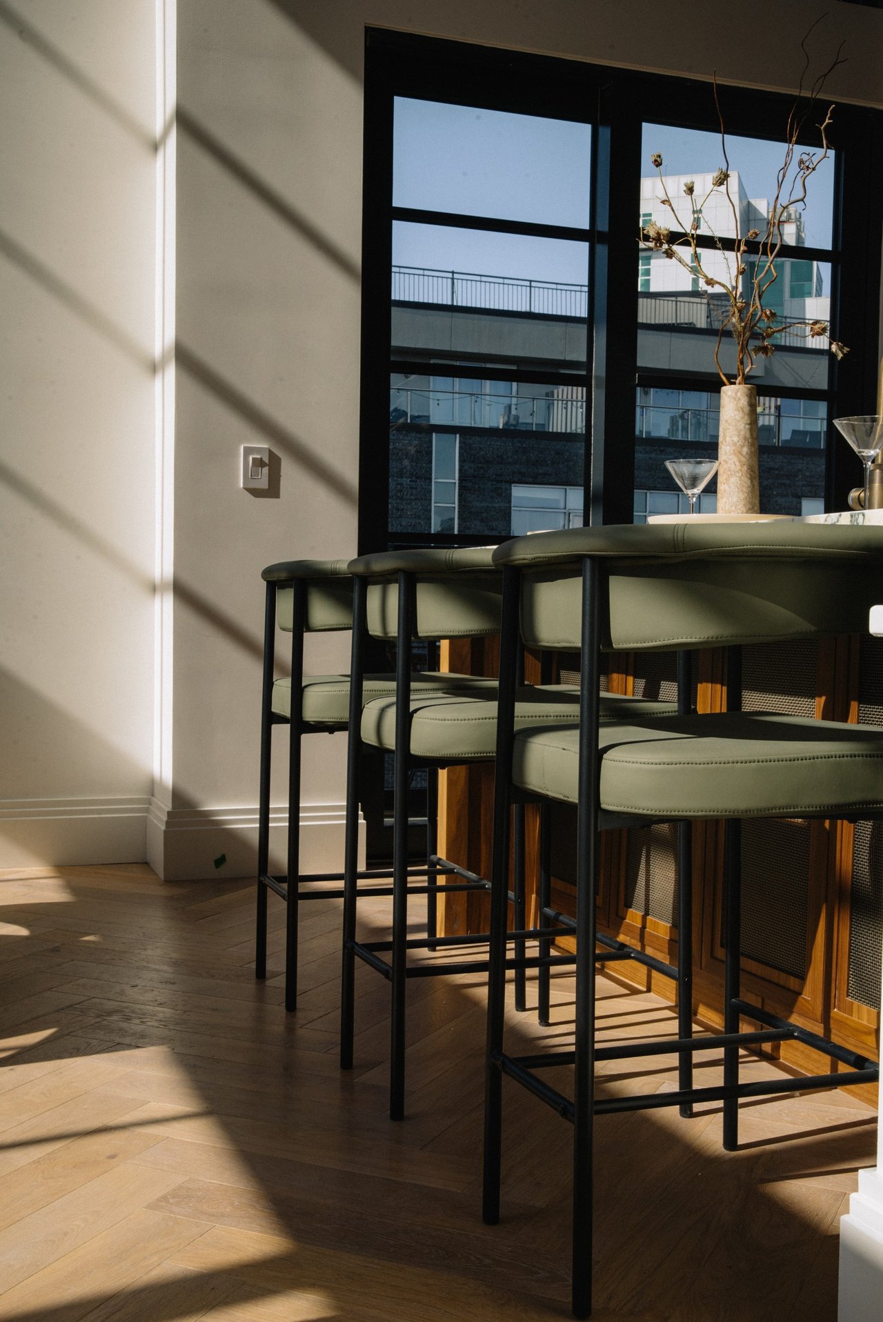 Kitchen counter with olive-green bar stools and large black-framed windows overlooking neighboring buildings.