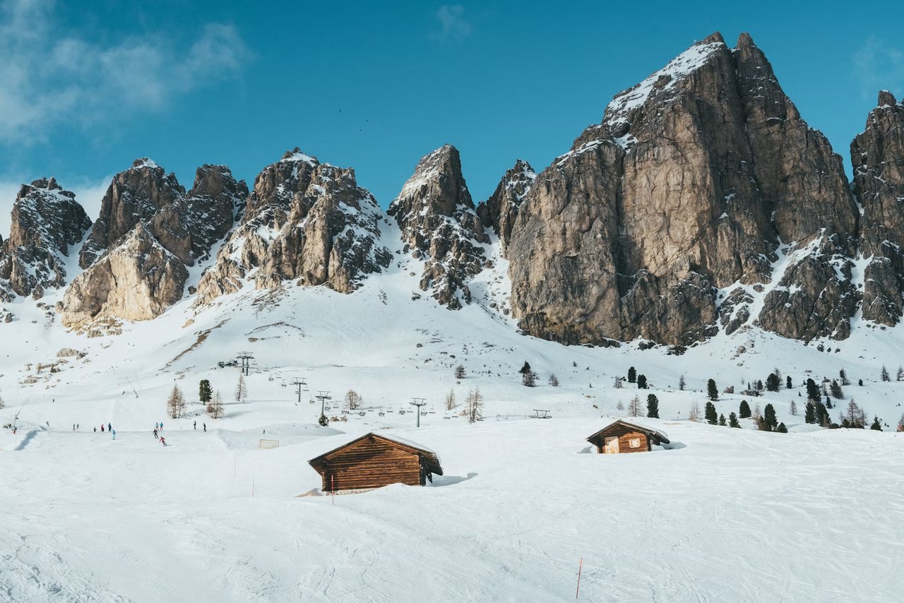 Skiers on a snowy slope beneath a jagged mountain ridge, near two wooden huts.