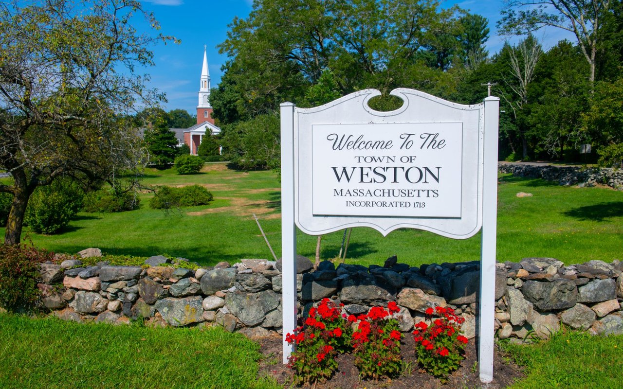 Weston, Massachusetts welcome sign beside a church steeple and red flowers.