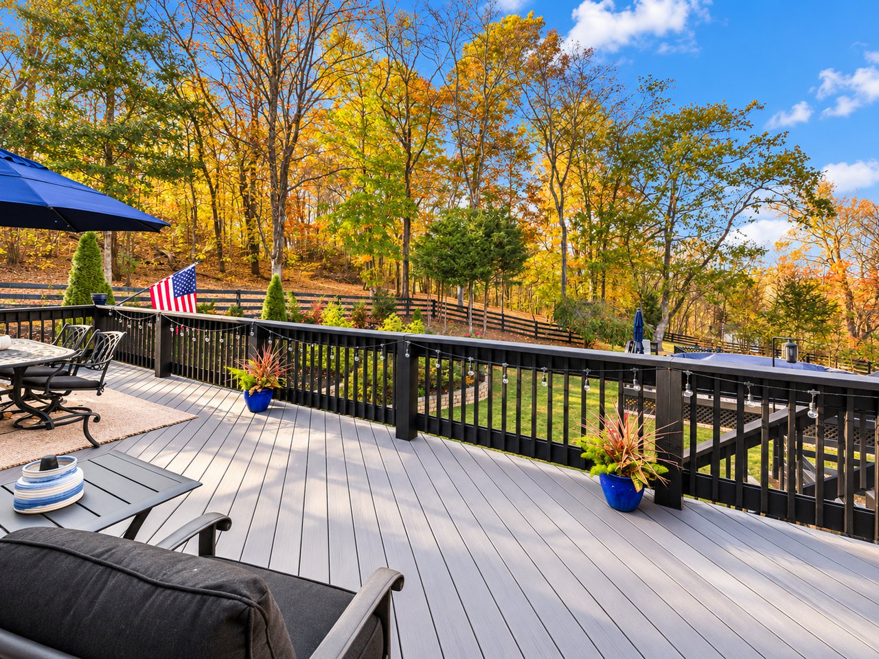 Furnished deck overlooking a fenced yard and autumn trees, with black railings and an American flag.
