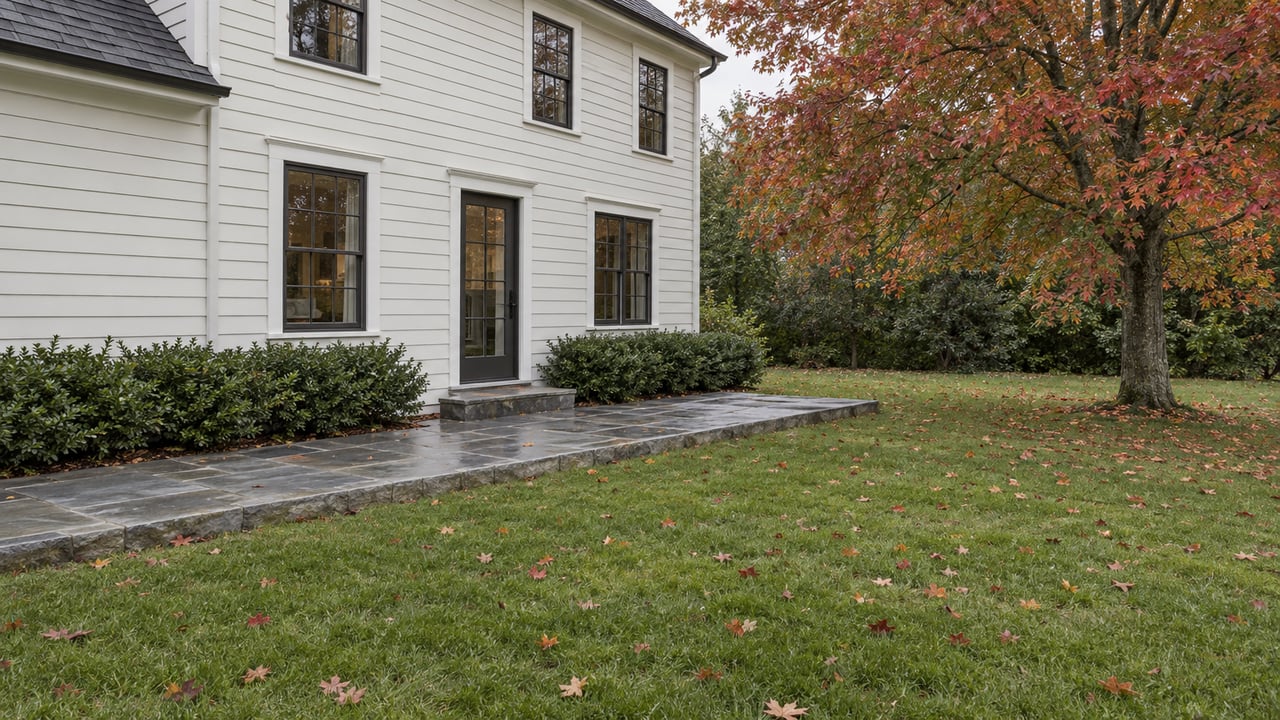 Off-white Colonial rear wall beside a bluestone terrace, deep lawn, and red maple with autumn leaves.