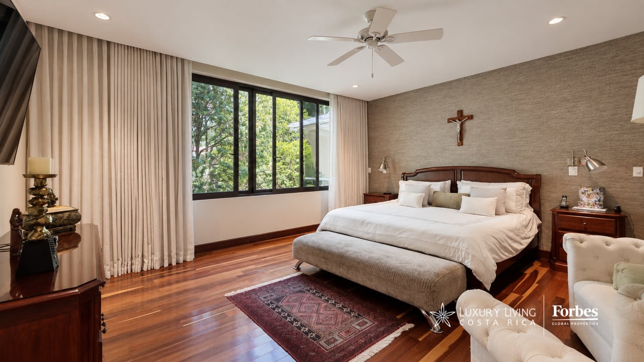 Bedroom with a large wooden bed, polished hardwood floors, and wide windows overlooking leafy trees.