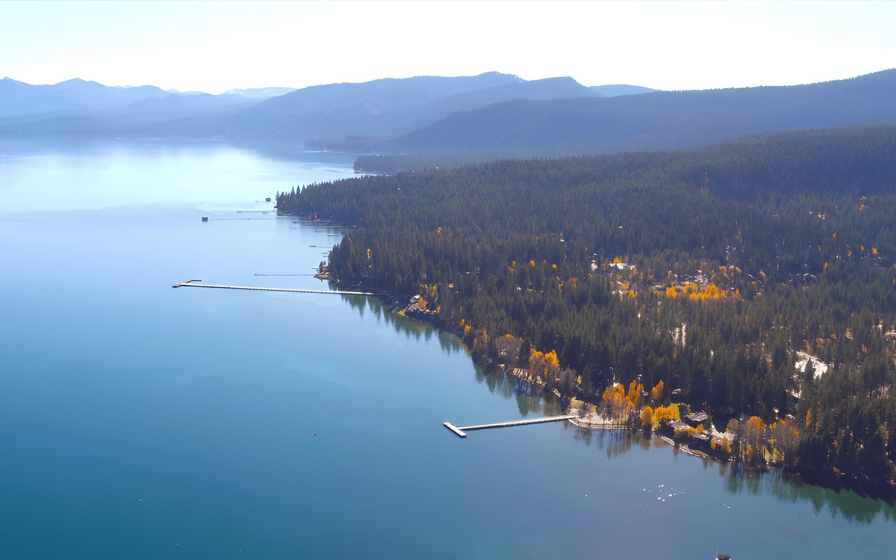 Aerial view of West Lake Tahoe’s forested shoreline, piers, and distant mountain ridges.