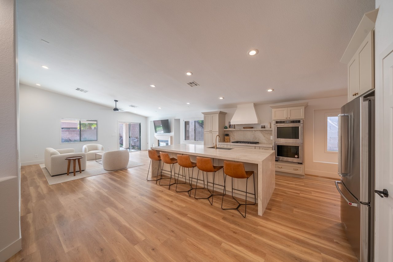 Open-plan kitchen with a stone island, orange stools, white cabinetry, and an adjoining living area.