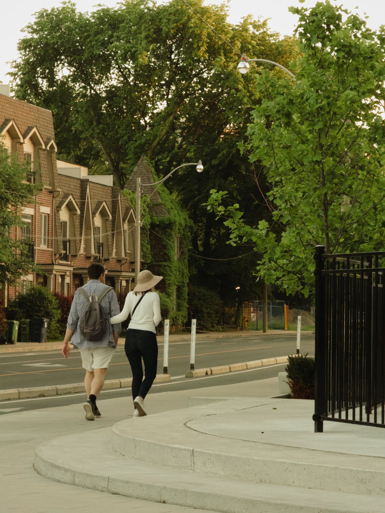 Two people walk beside brick townhomes on a leafy sidewalk.