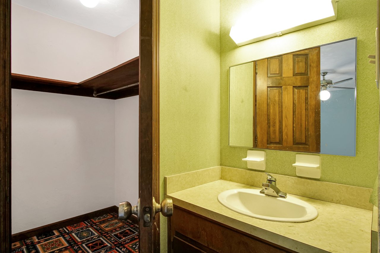 A small bathroom features green walls, a beige countertop with a sink, and a large mirror reflecting a wooden door. The adjacent room has a colorful patterned carpet.