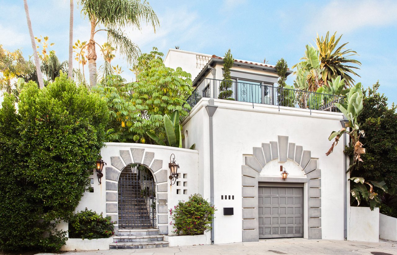 White stucco home with an arched entry gate, gray garage door, and tropical landscaping.