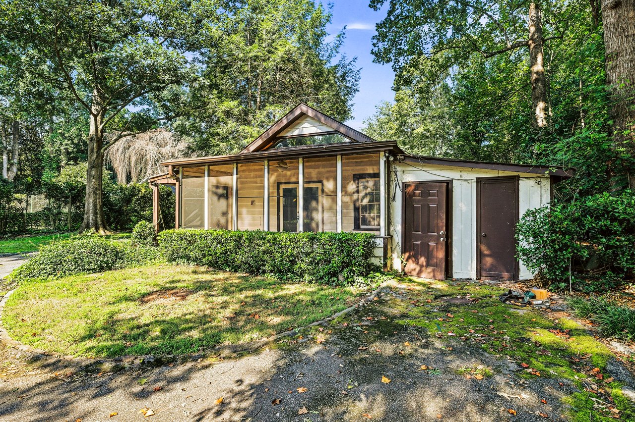 Single-story house with a screened porch and triangular gable window among mature trees.