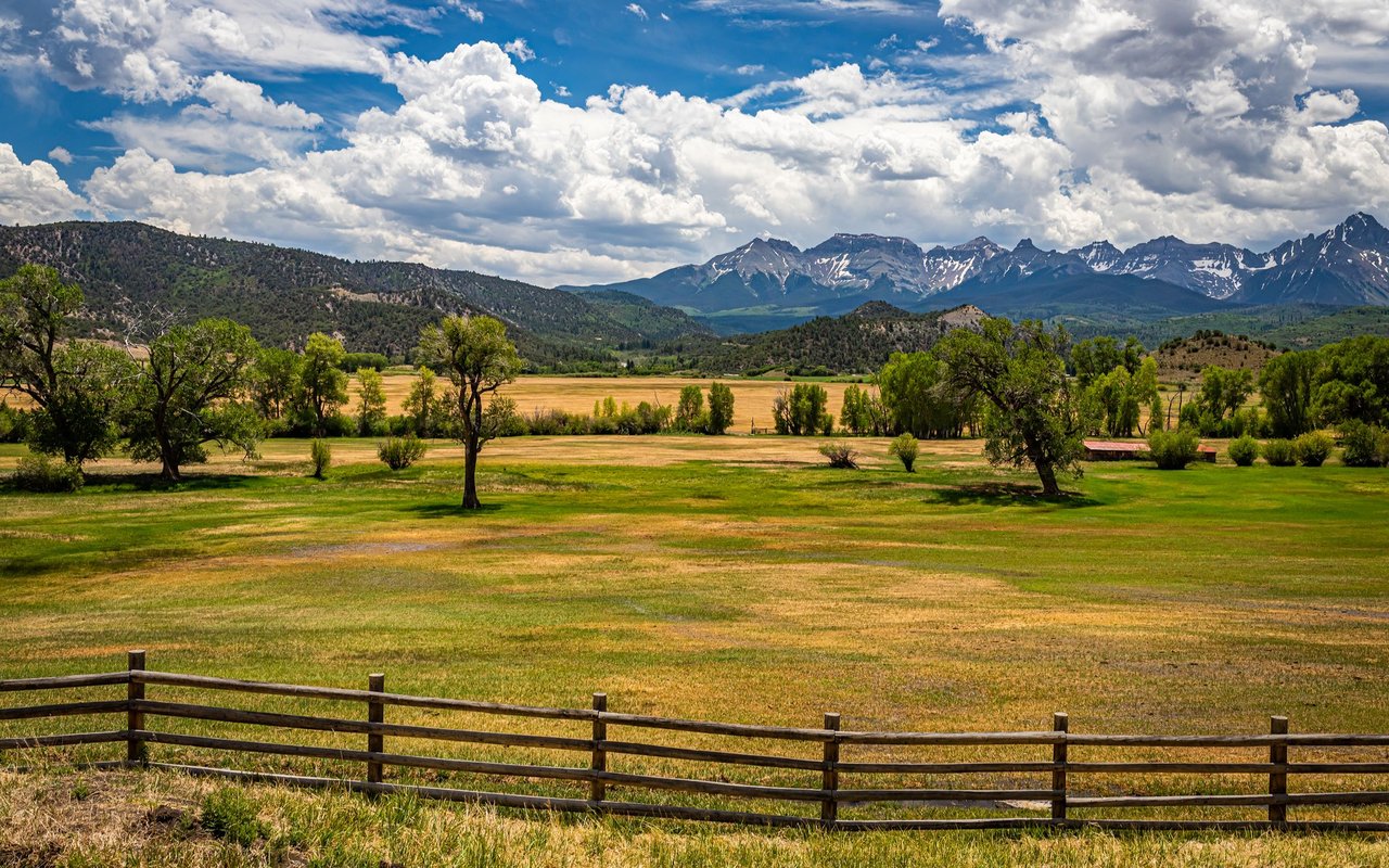 Open meadow and wooden fence with forested foothills and snow-streaked mountains beyond.