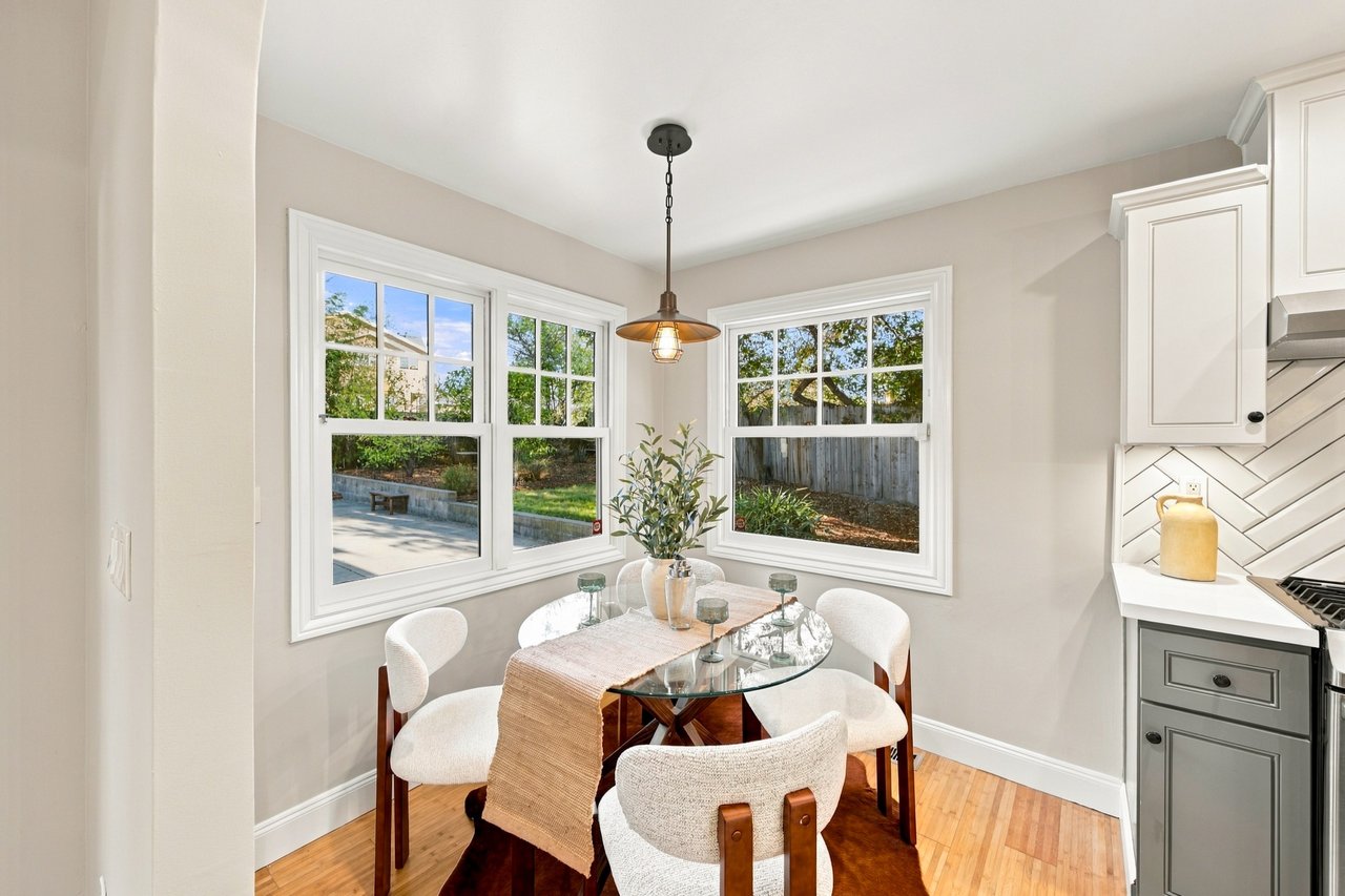 Dining nook with a glass table and upholstered chairs beside three windows overlooking a fenced yard.