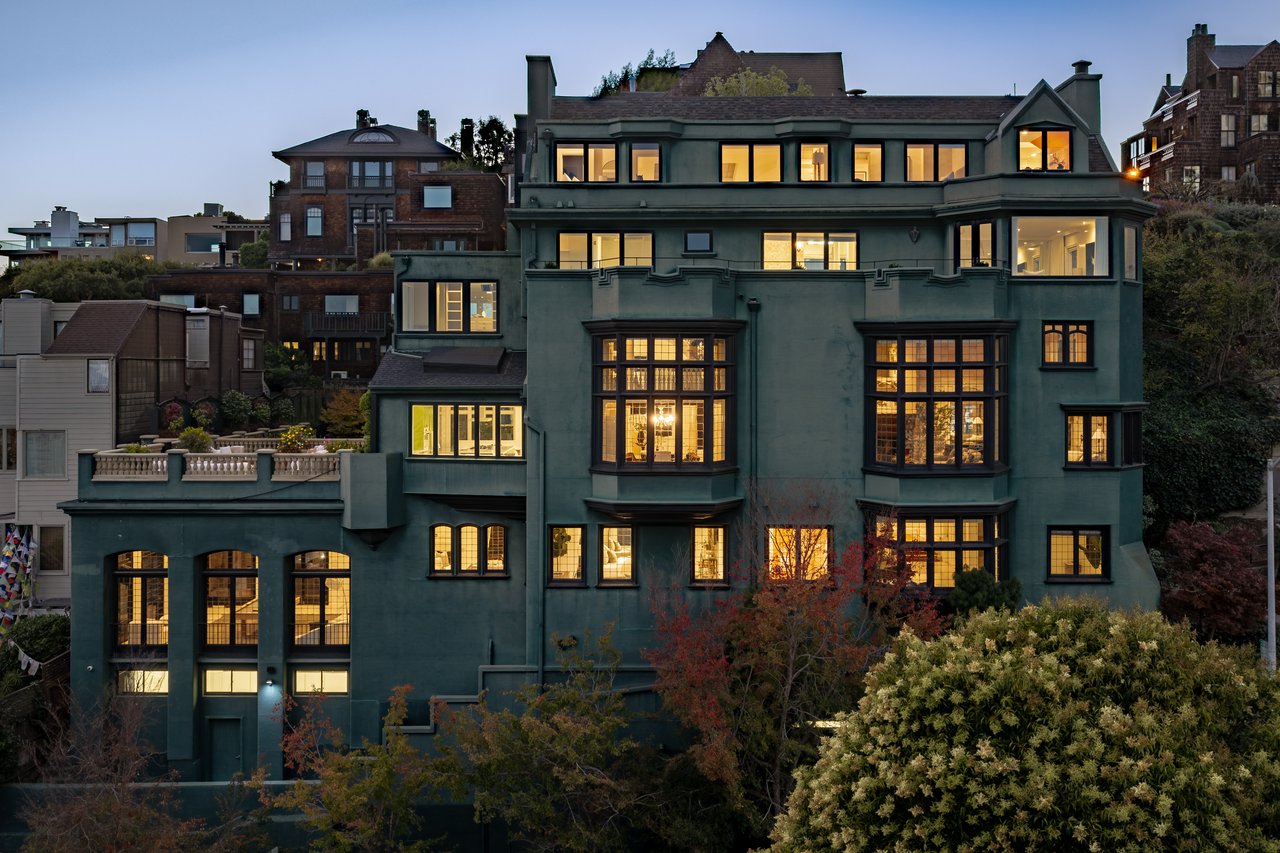 Multi-story dark green home with warmly lit windows, bay windows, and neighboring buildings at dusk.