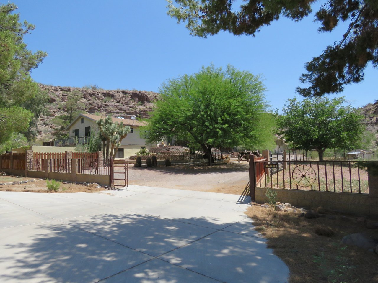 Desert home behind a decorative metal-and-wood fence, with rocky hills and mature trees.