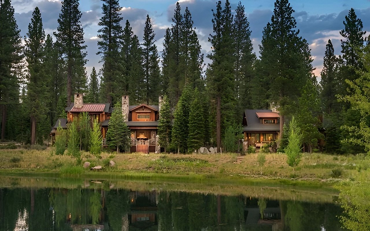 Wood-and-stone homes among pines beside a pond, reflected in the water at dusk.
