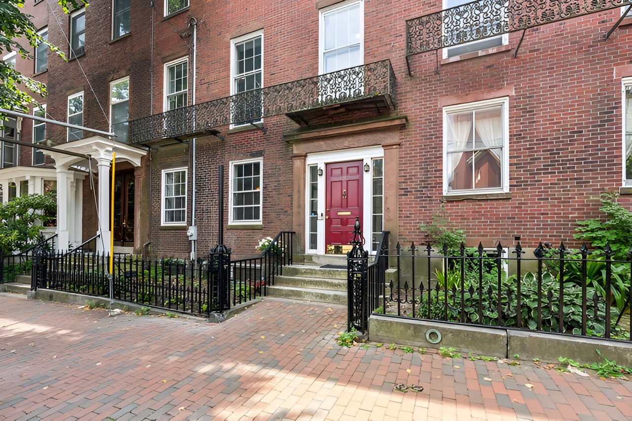 Red-brick building with a red entry door, ornate iron balconies, and a fenced brick-front garden.