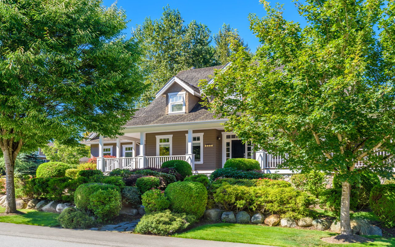 a single story house with a brown exterior and a white front door with a porch, surrounded by trees and bushes