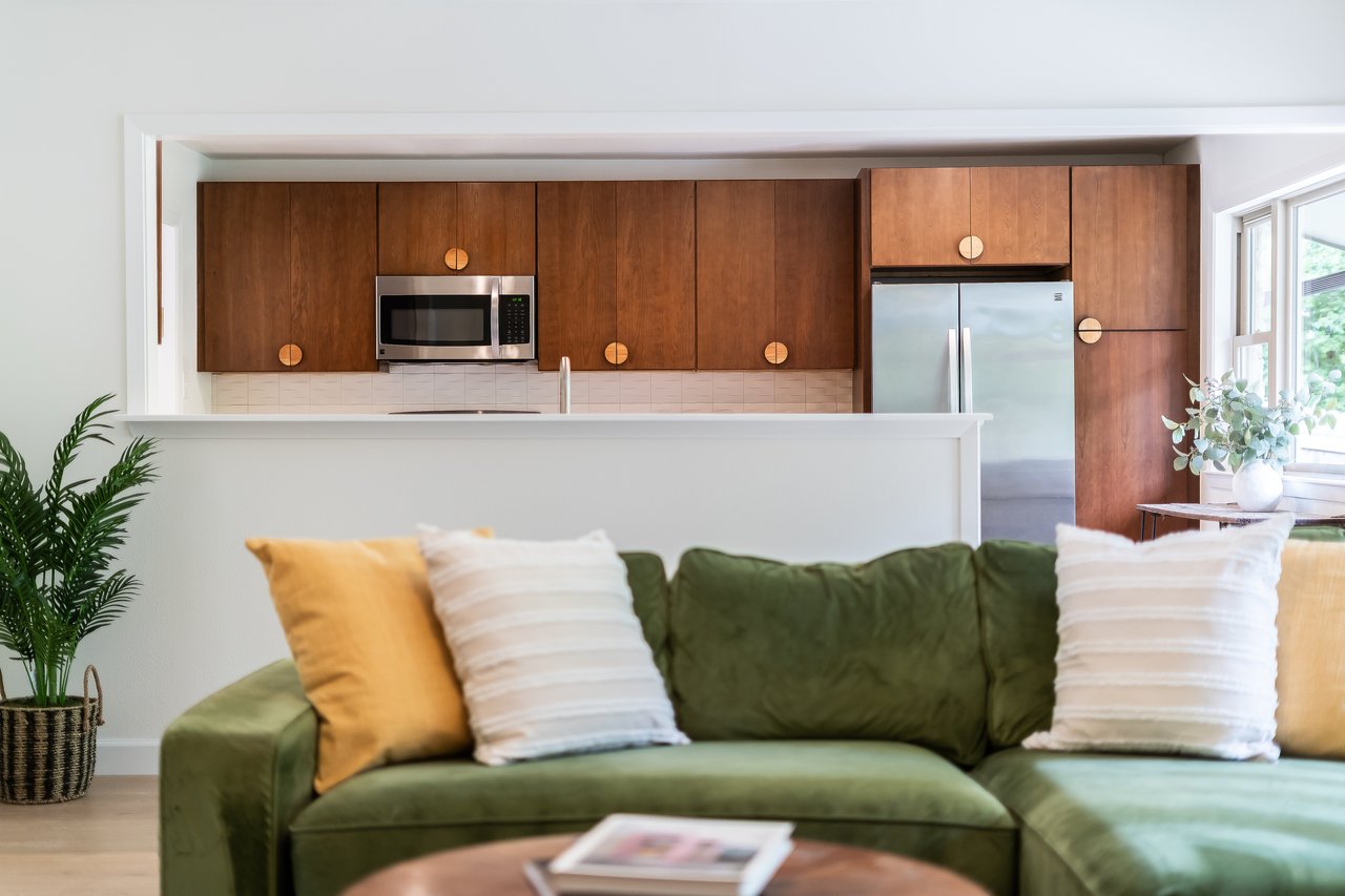 Green sectional sofa faces a kitchen with warm wood cabinets and stainless-steel appliances.